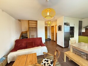 A modern living room with a sofa, a dining table, and a kitchen area with a refrigerator and microwave. - Chalet cosy à Risoul village • Terrasse & vue vallée (Risoul)