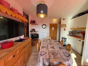 A compact living space with a TV, wooden furniture, a dining table, and a kitchenette.