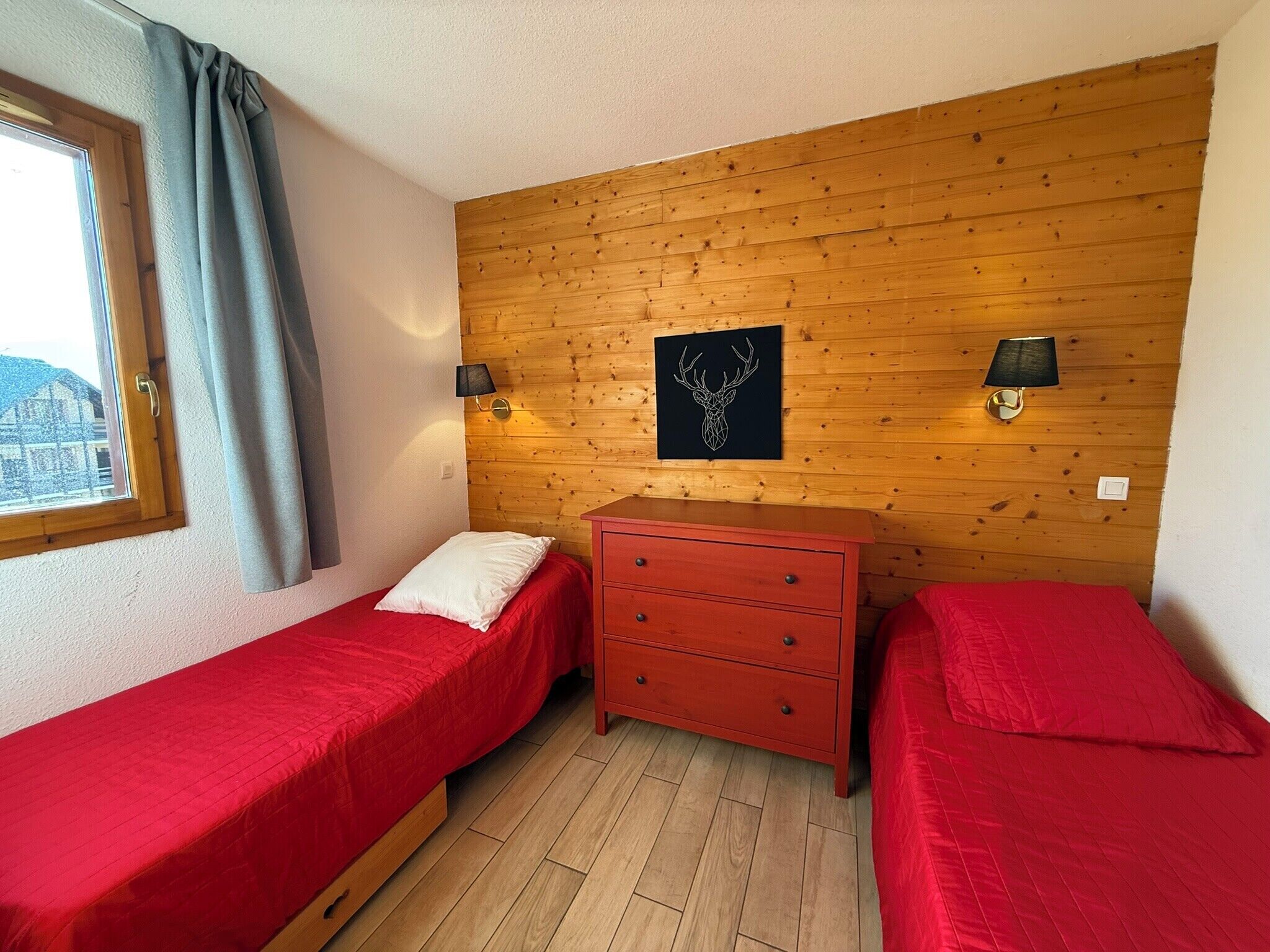 Habitació petita i acollidora amb dos llits individuals, un panell de paret de fusta, un calaixer vermell i dues làmpades penjades a la paret.