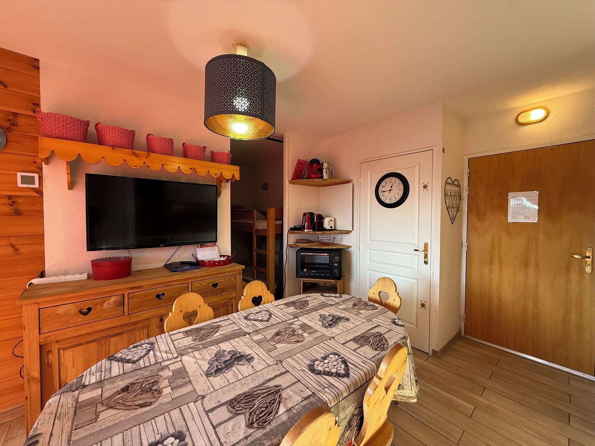 Um quarto aconchegante com TV, móveis de madeira, toalha de mesa estampada, micro-ondas e um relógio na parede.