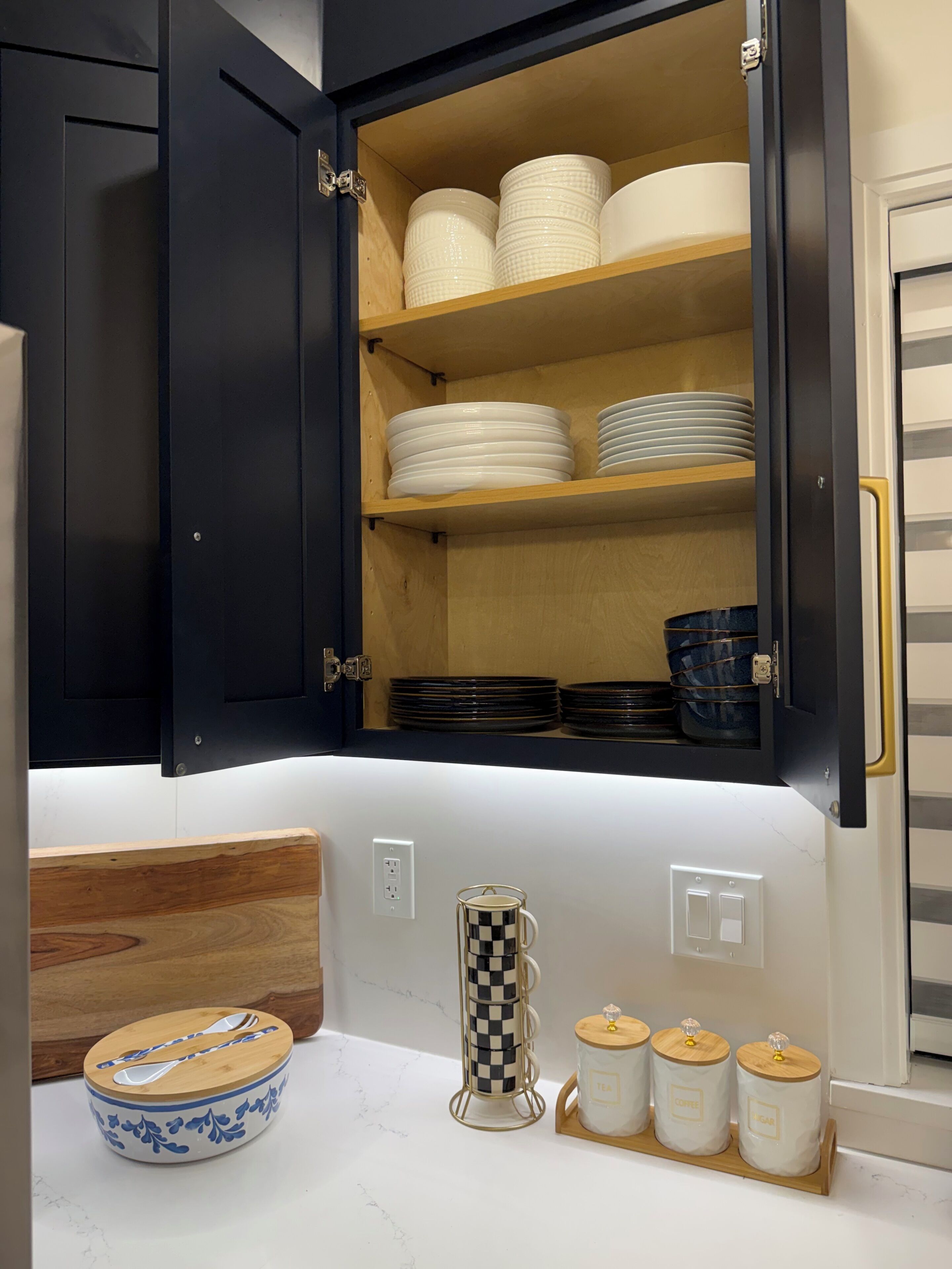 Une cuisine avec des meubles de couleur sombre, un plan de travail en bois, un bol avec un couvercle, un porte-tasses à carreaux et trois bocaux étiquetés « thé », « café » et « pain ».