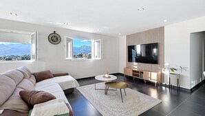 Ein modernes Wohnzimmer mit einer Couch, einem Fernseher und Blick auf Berge durch die Fenster.
