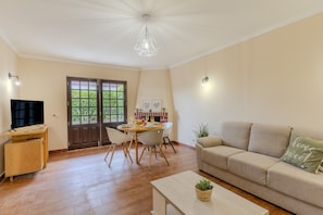 Ein Wohnzimmer mit Sofa, Couchtisch, Fernseher, Essbereich mit Tisch und Stühlen sowie Küchenzeile mit Herd und Schränken.