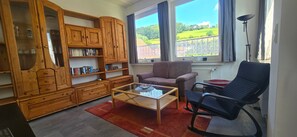 A living room with a wooden cabinet, a sofa, a chair, and a view of a green landscape through large windows. - Apartment 'FeWo Meyer in Hüddingen' with Private Garden and Wi-Fi (Bad Wildungen)