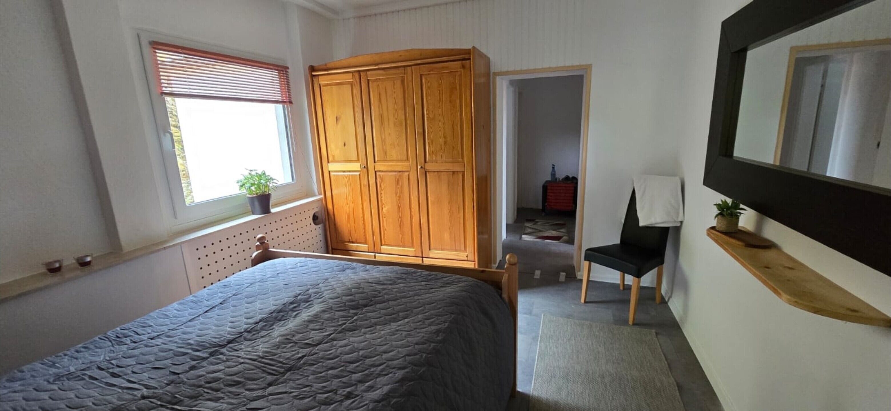 Un dormitorio con cama, armario de madera, silla, escritorio, espejo y ventana con persianas.
