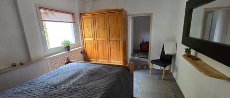 Un dormitorio con cama, armario de madera, silla, escritorio, espejo y ventana con persianas.