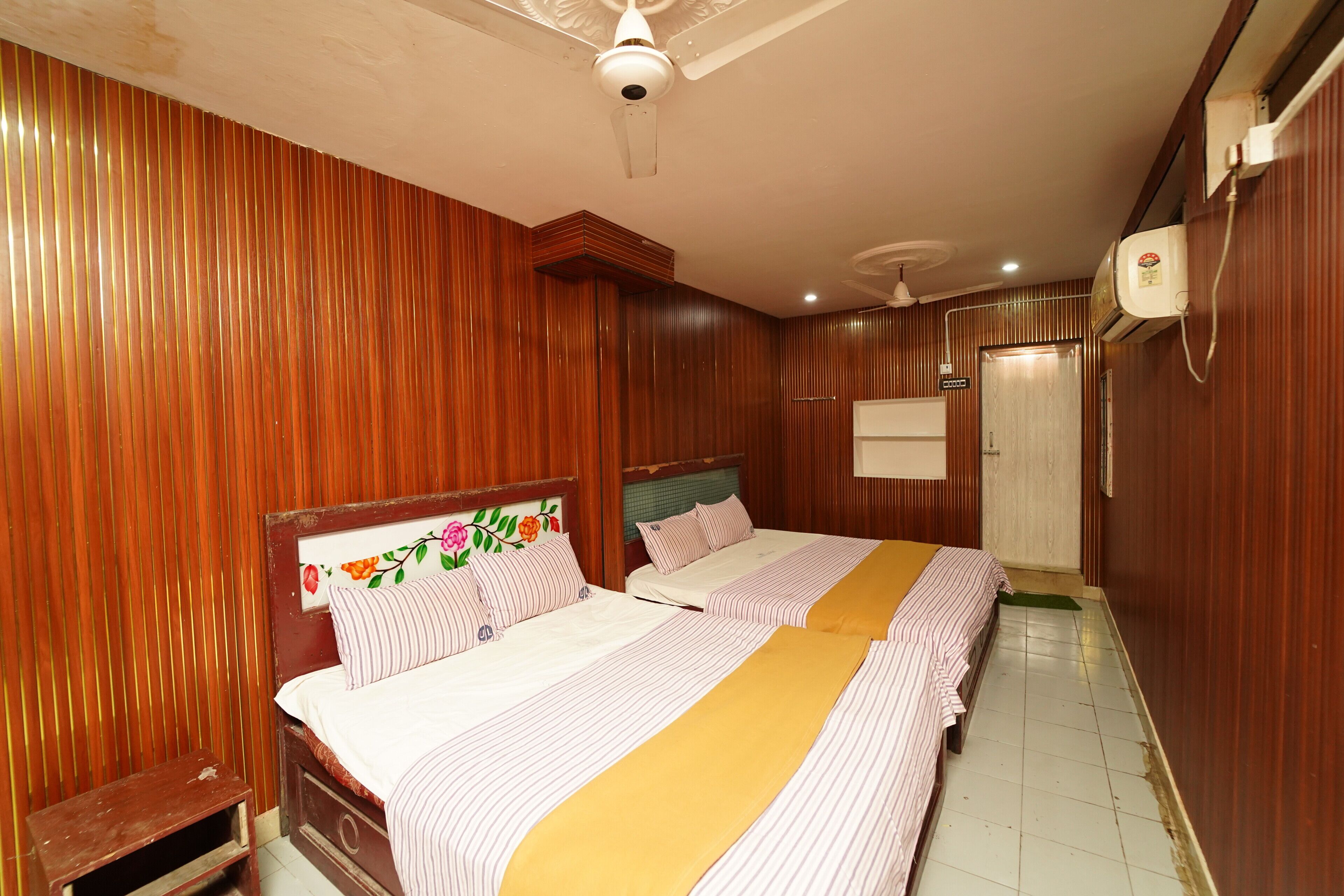 Habitación de hotel con dos camas, paneles de madera, ventilador de techo, aire acondicionado y un taburete de madera.
