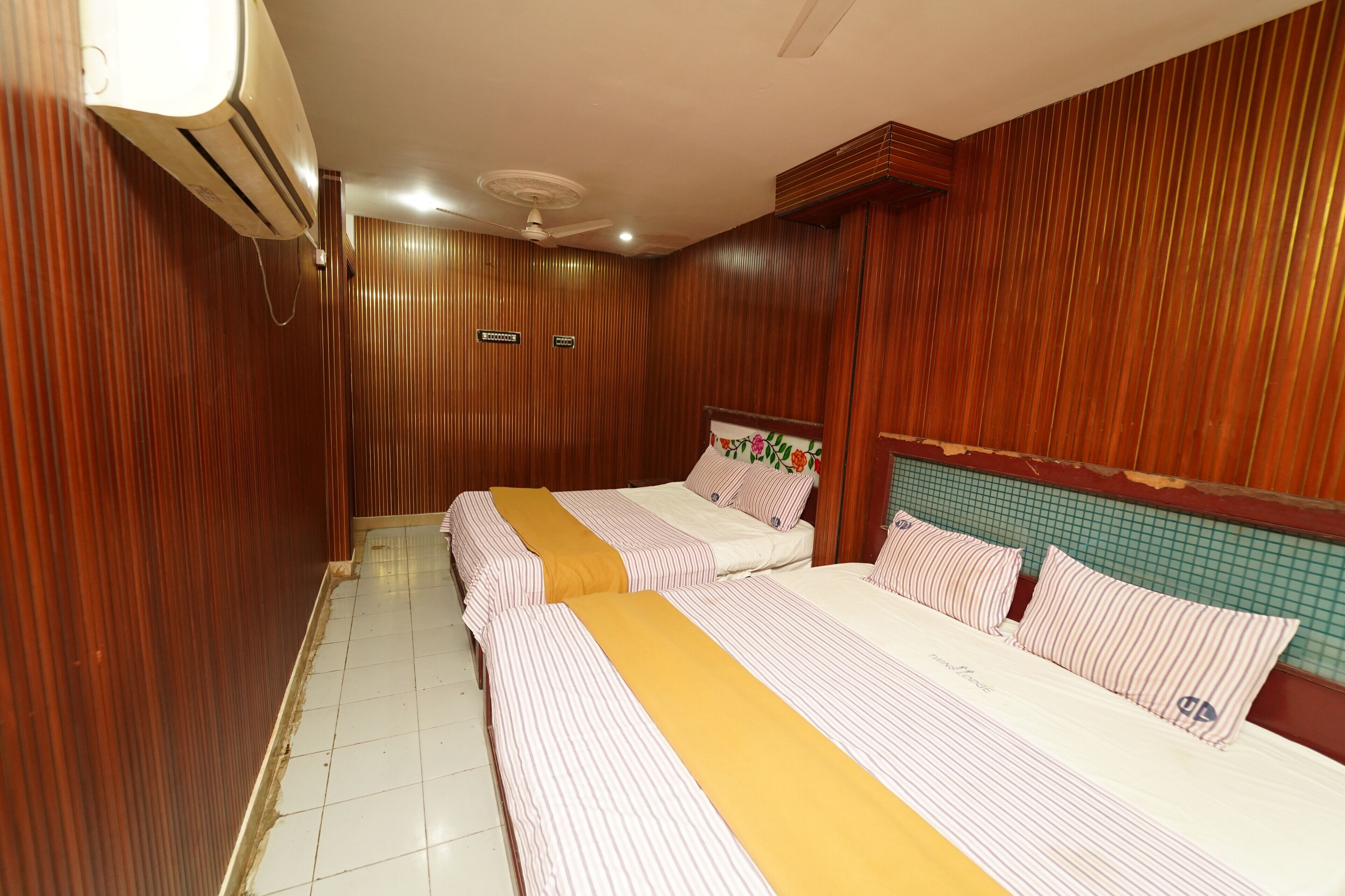 Quarto de hotel com duas camas, painéis de madeira, piso de azulejos, ar condicionado e ventoinha no teto.