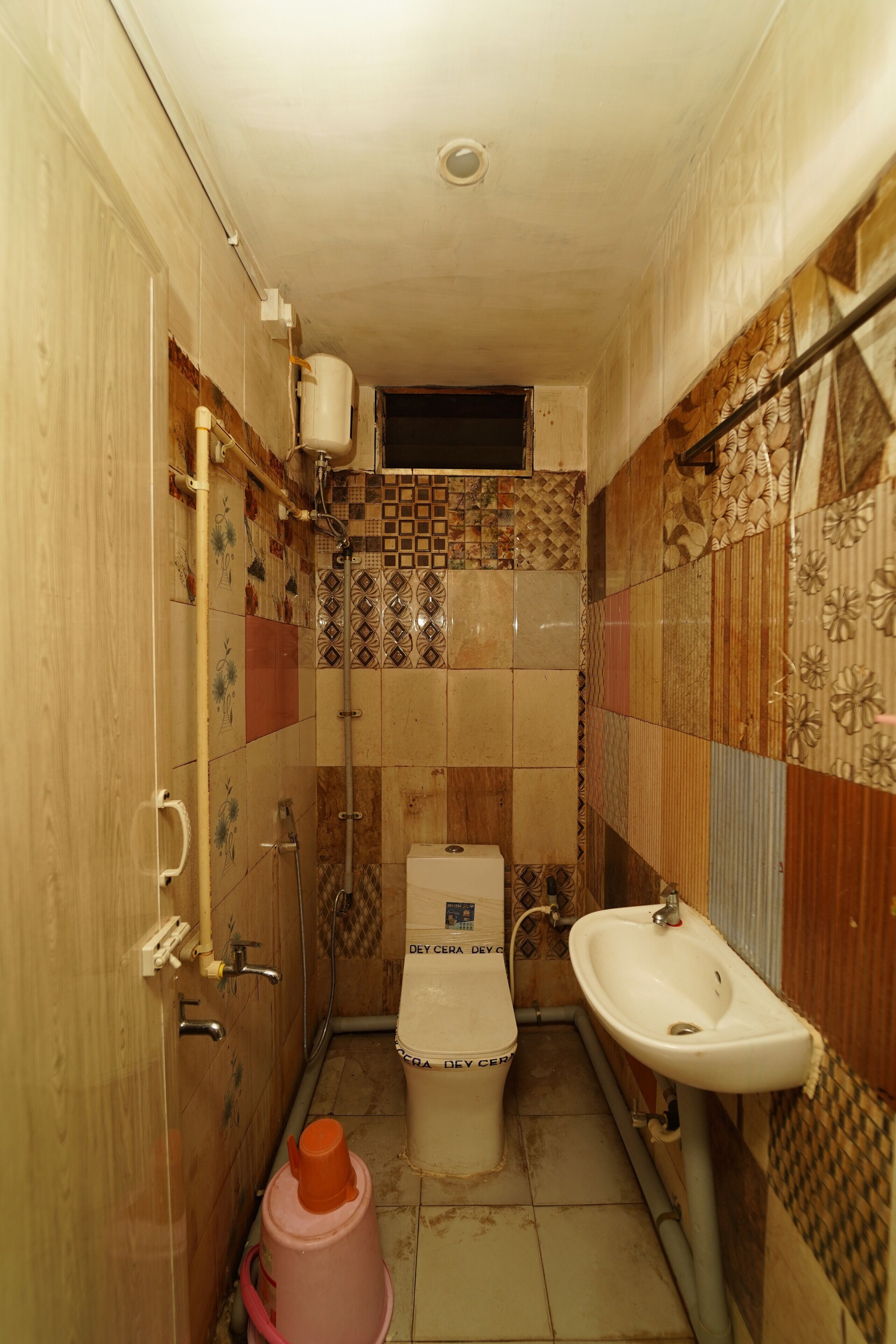 A small, narrow bathroom with a toilet, sink, and shower. The walls are tiled with various patterns and colors.