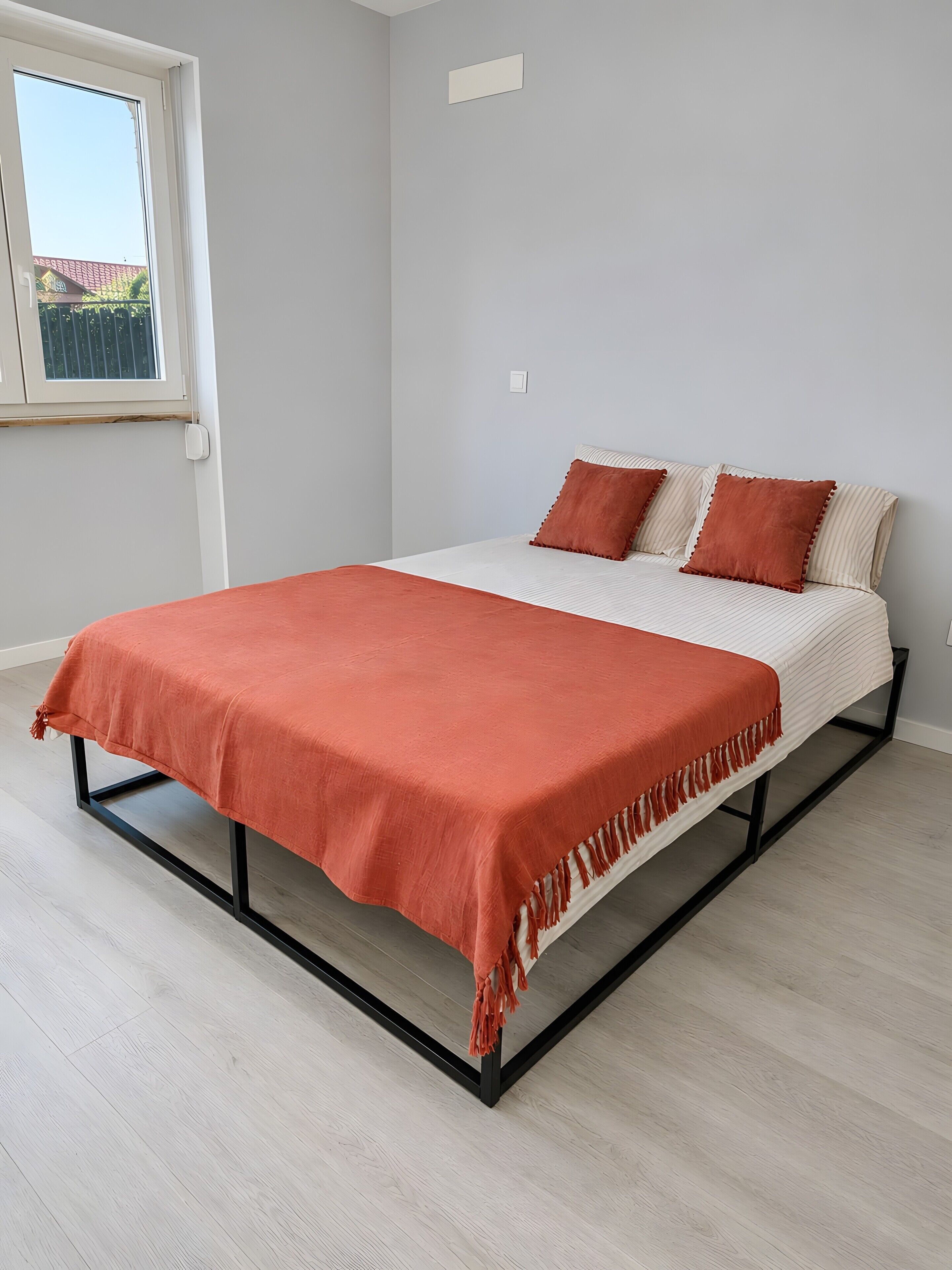 Una cama con una colcha y almohadas rojas, una ventana con vistas y un piso de color claro.