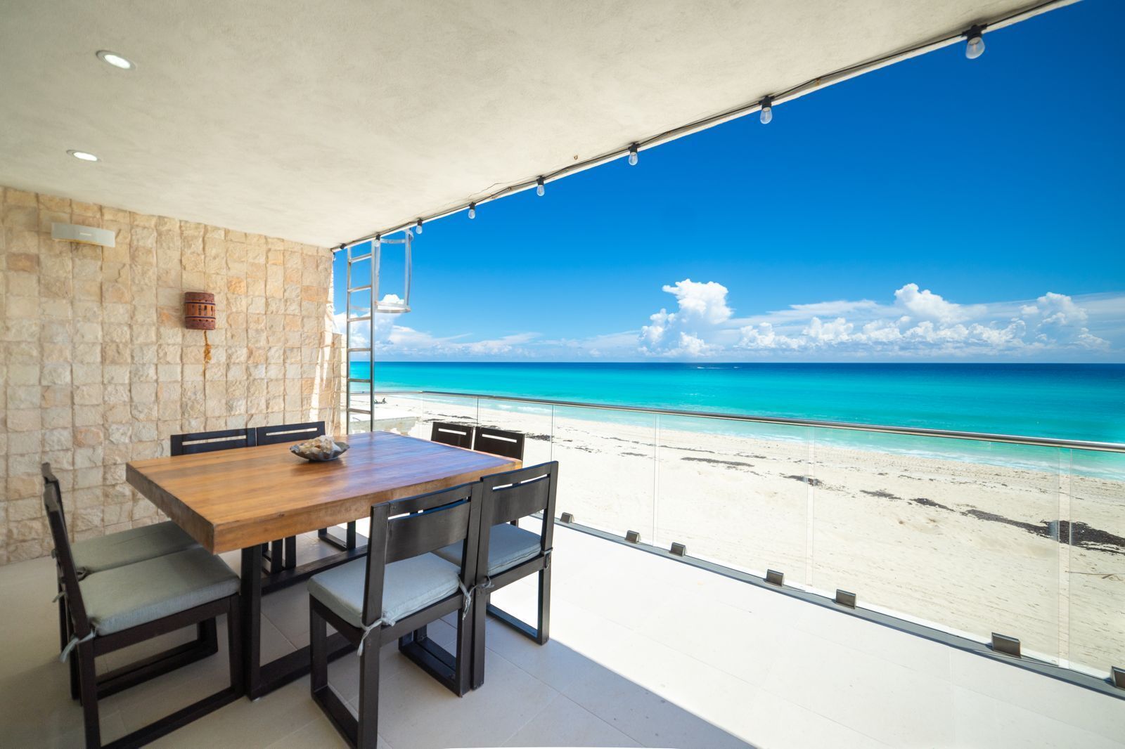A balcony with a wooden table and chairs, a stone wall, and a view of a beach and ocean.