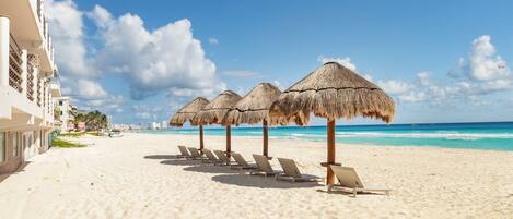 A beach with thatched umbrellas and sun loungers, a clear blue sky, and a building on the left.