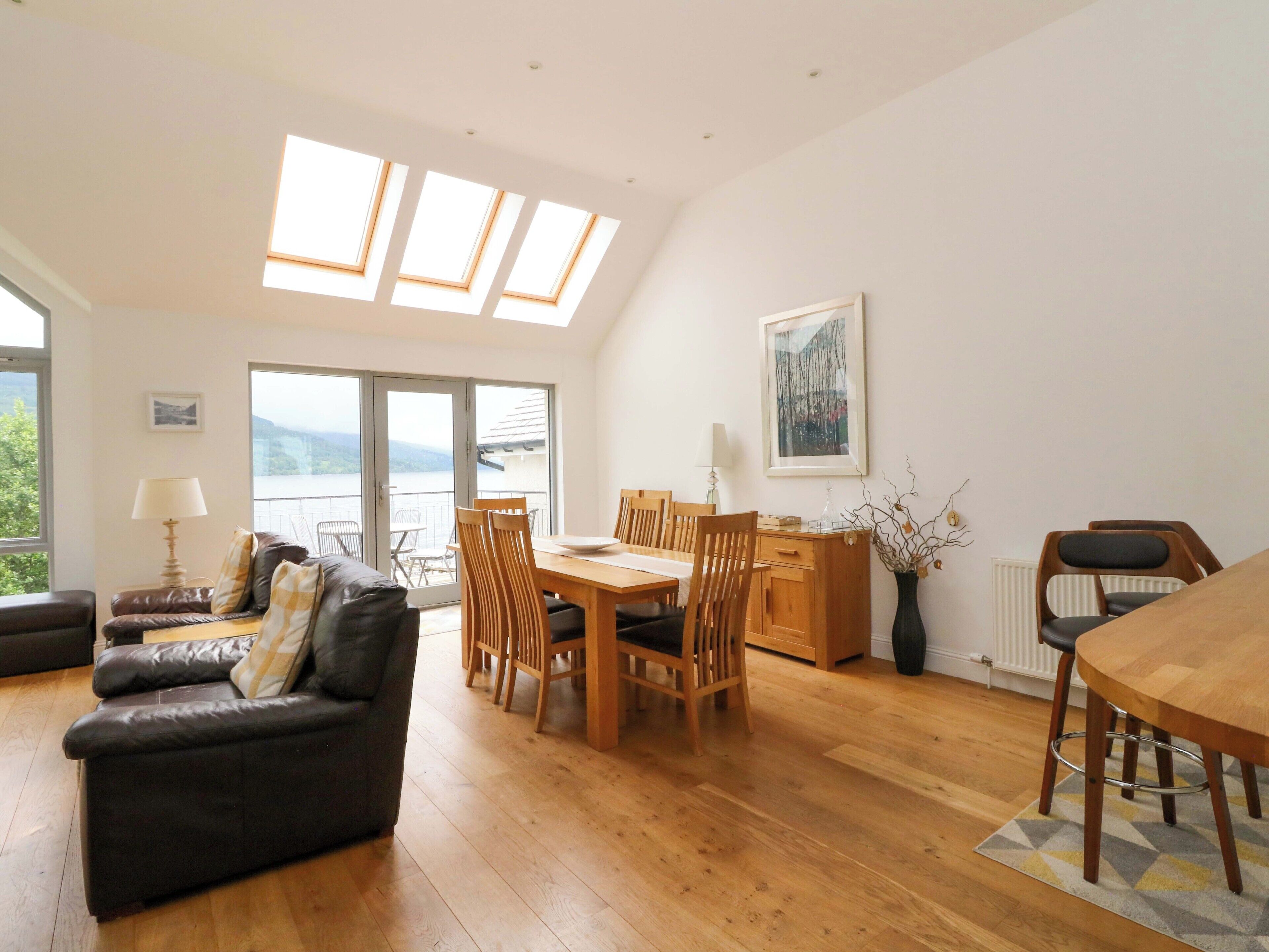 A modern living room with a wooden dining table, a sofa, and a view of a body of water through large windows.