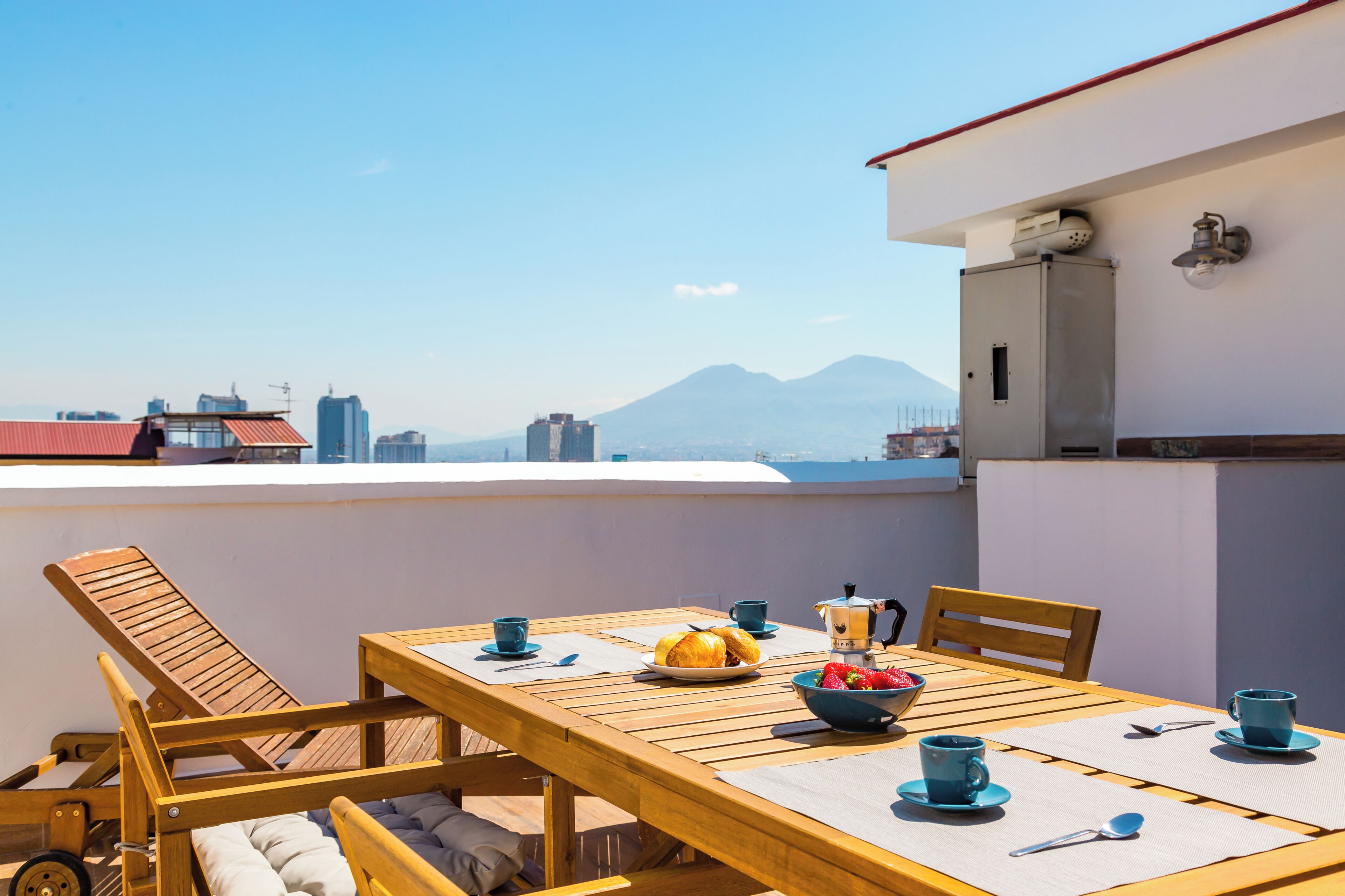 A wooden table with a view of a mountain, a bowl of fruit, and blue cups on a balcony.
