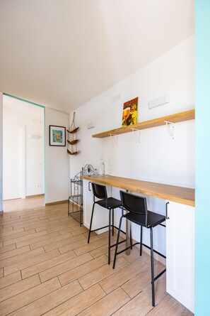 Ein Zimmer mit Holzboden, einer weißen Tür, einem an der Wand befestigten Regal mit einem Bild, zwei schwarzen Stühlen und einem Tisch.