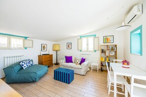 Ein Zimmer mit einer Couch, einem Esstisch, einer Lampe, einem Heizkörper und einem Bücherregal.