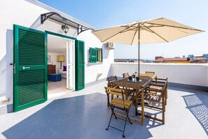 Eine Terrasse im Freien mit Tisch und Stühlen, einem großen Sonnenschirm und grünen Fensterläden an einem weißen Gebäude.