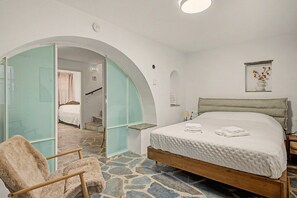 Een slaapkamer met een vloer met steenmotief, een bed met een witte dekbedovertrek, een houten stoel en een glazen deur naar een badkamer.