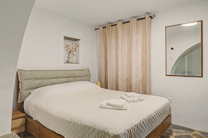 Un dormitori amb un llit, una taula de nit, una làmpada, un mirall i una finestra amb cortines.