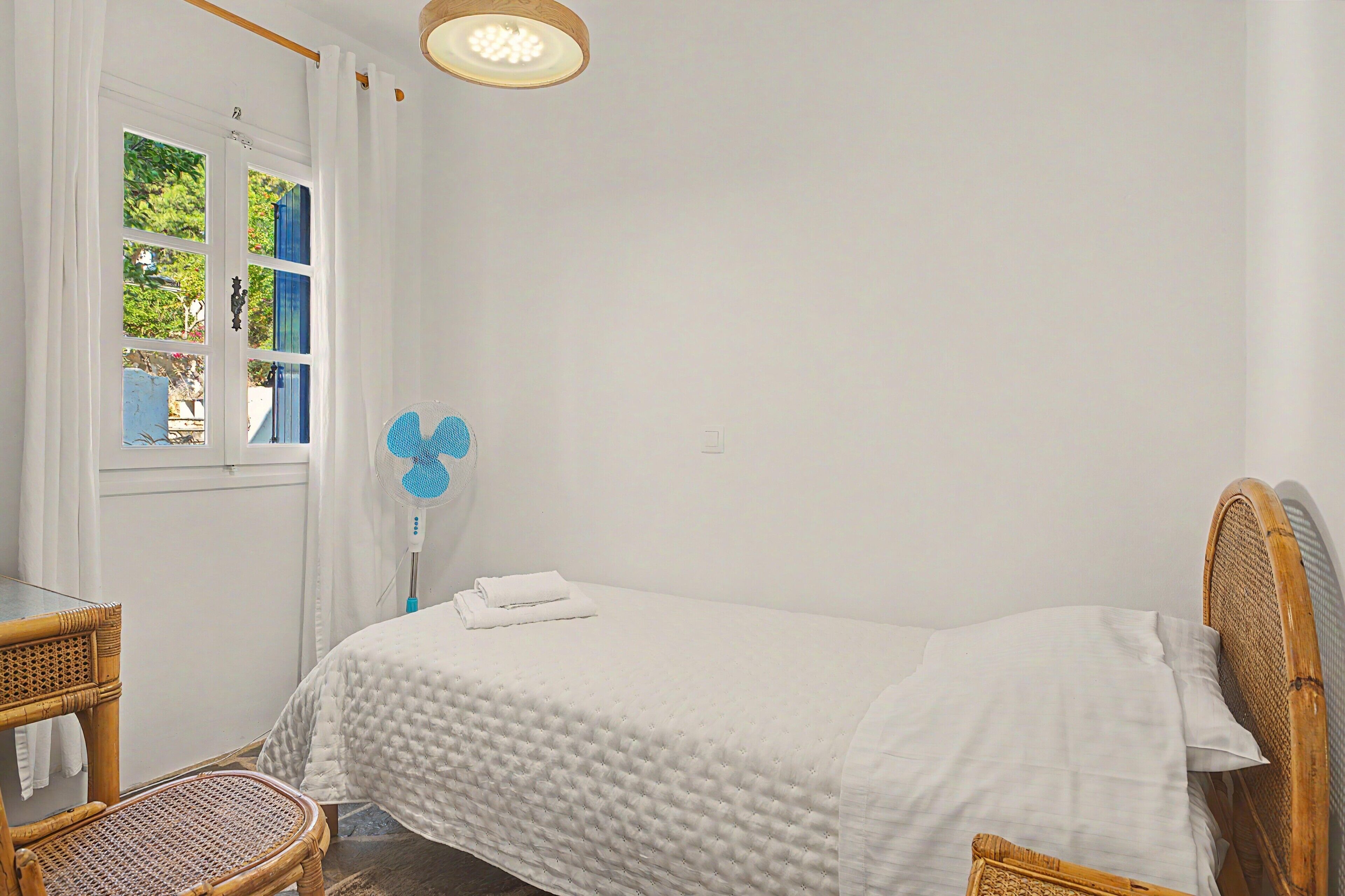 A single bed with a white quilt, a bedside table, a chair, and a window with curtains.