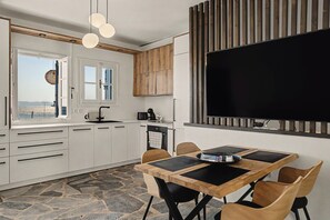 Eine moderne Küche mit weißen Schränken, Steinboden, einer Essecke mit Holztisch und -stühlen sowie einem großen, an der Wand montierten schwarzen Fernseher.