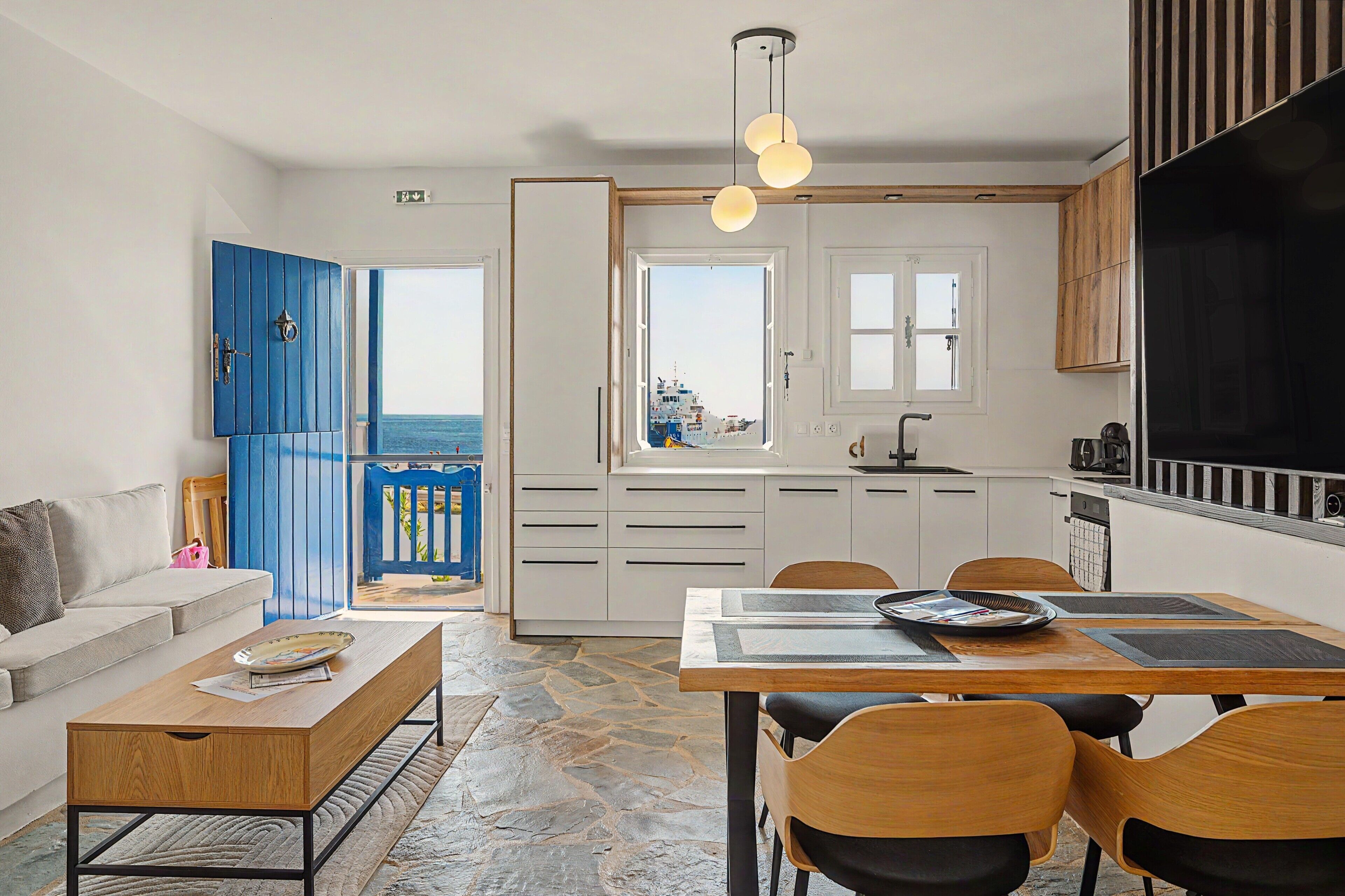 A modern kitchen with white cabinets, a black countertop, and a view of the sea through the open door.