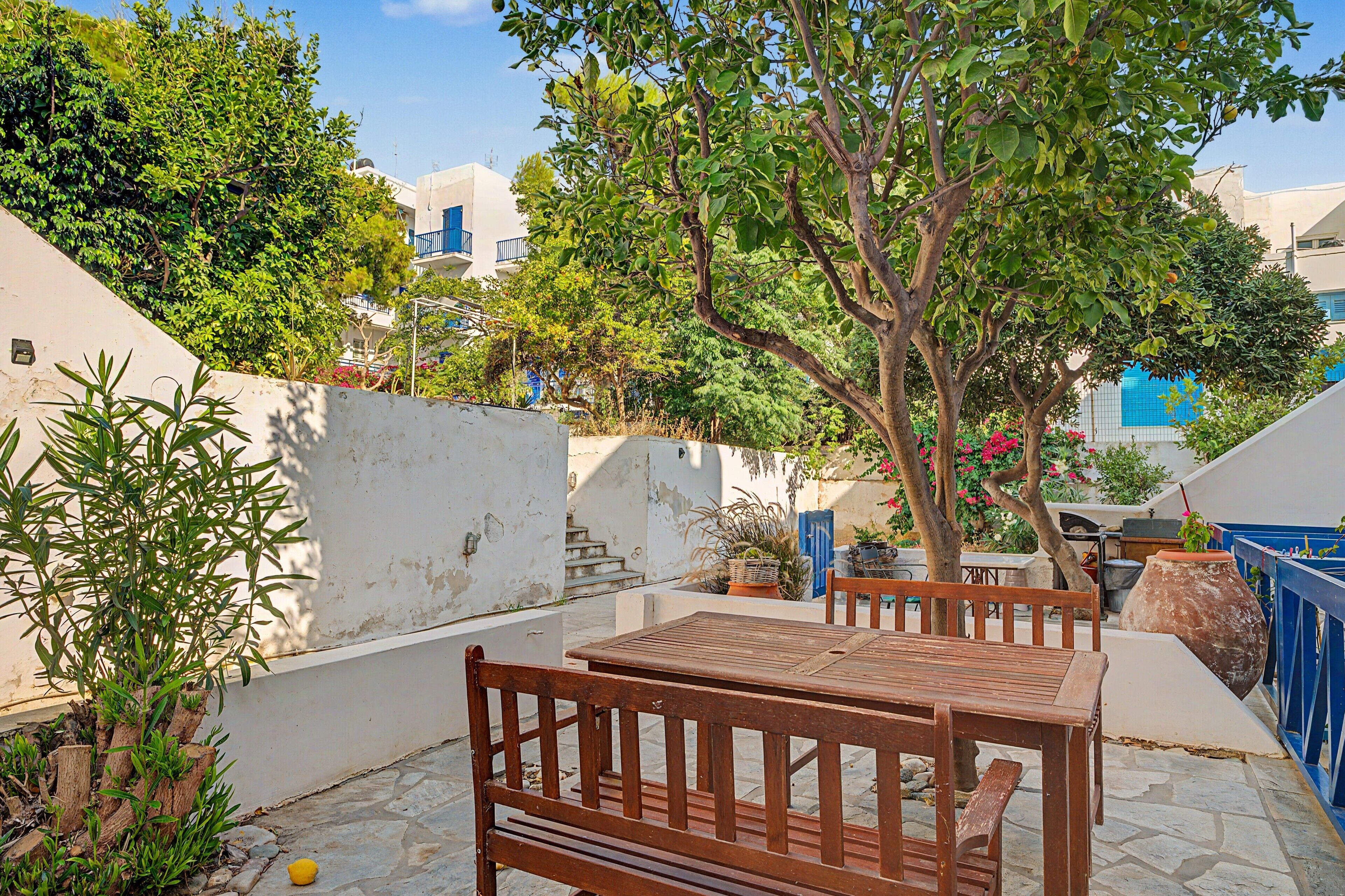 An outdoor seating area with a table and benches, surrounded by greenery and a white wall.
