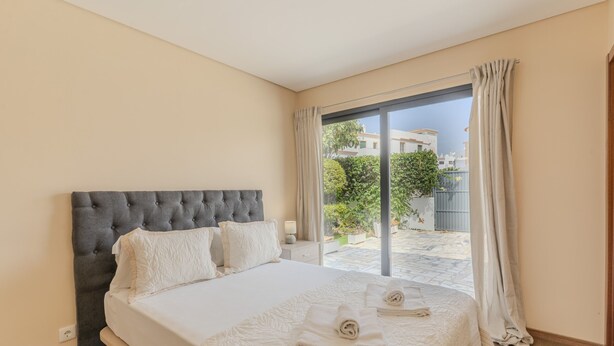 A hotel room with a large bed, a grey upholstered headboard, and a view of a courtyard through a sliding glass door. - #390 New Town Apartment with Shared Pool (Albufeira)