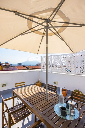 A wooden table with a metal umbrella, a wicker basket, and a potted plant on a rooftop terrace with a view of a cityscape. - Viewsuvio Family Terrace (Naples)