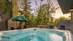 Ein Whirlpool mit einem grünen Sonnenschirm, umgeben von Bäumen und einer Terrasse mit Stühlen.