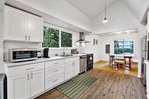 A modern kitchen with white cabinetry, a built-in oven, a microwave, and a coffee maker. There is a dining area with a table and chairs, and a window providing a view of the outdoors. - The Dale Home (Seattle)