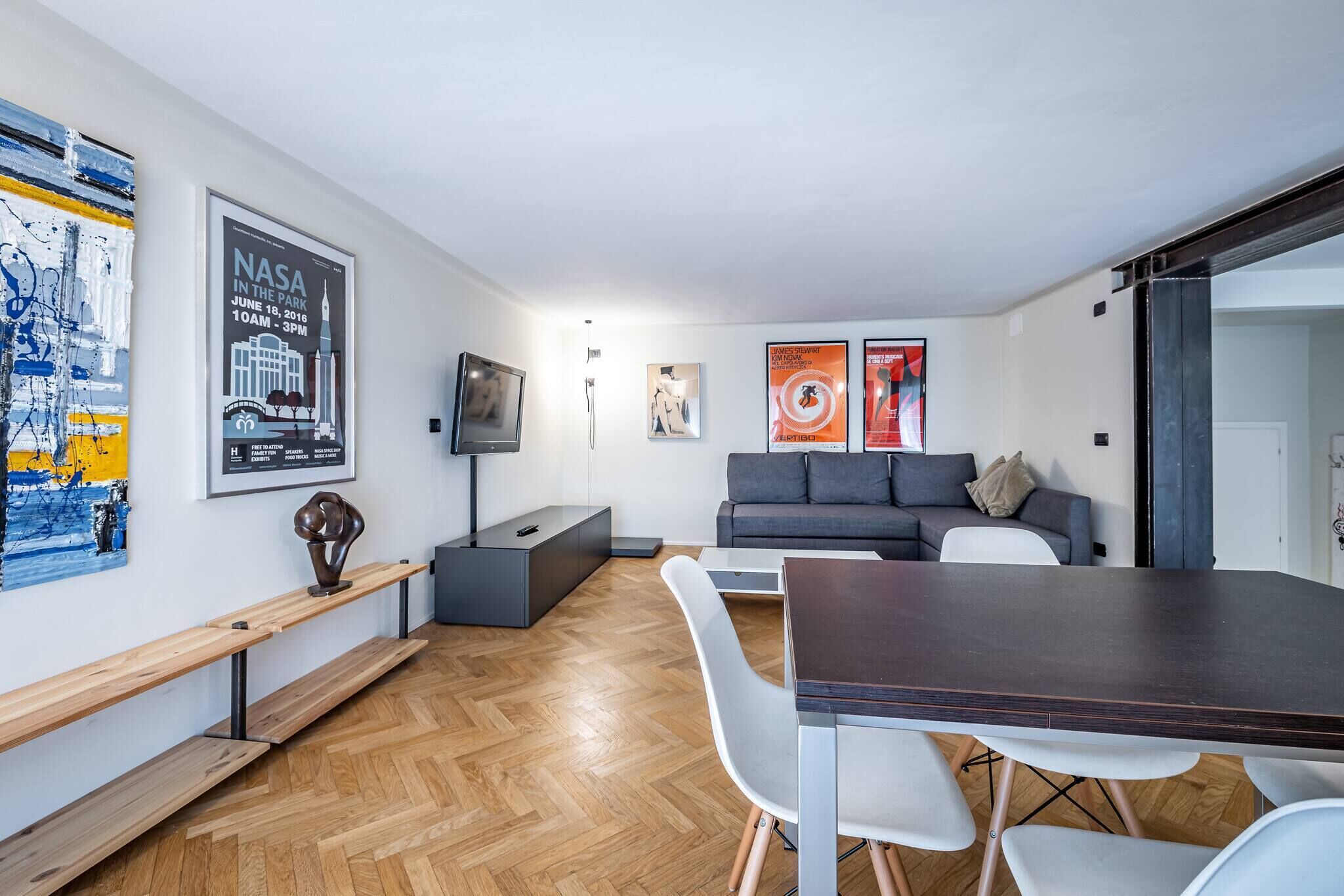 Un salon moderne avec un parquet en bois disposé en chevrons, une table à manger en bois foncé, des chaises blanches, un canapé gris et un téléviseur posé sur un meuble.