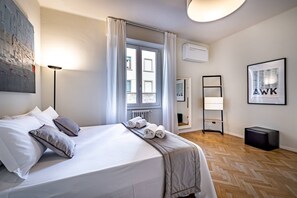 Une chambre d’hôtel moderne avec un lit soigneusement fait, une grande fenêtre avec des rideaux, une lampe sur pied, un pouf noir et un tableau encadré au mur.