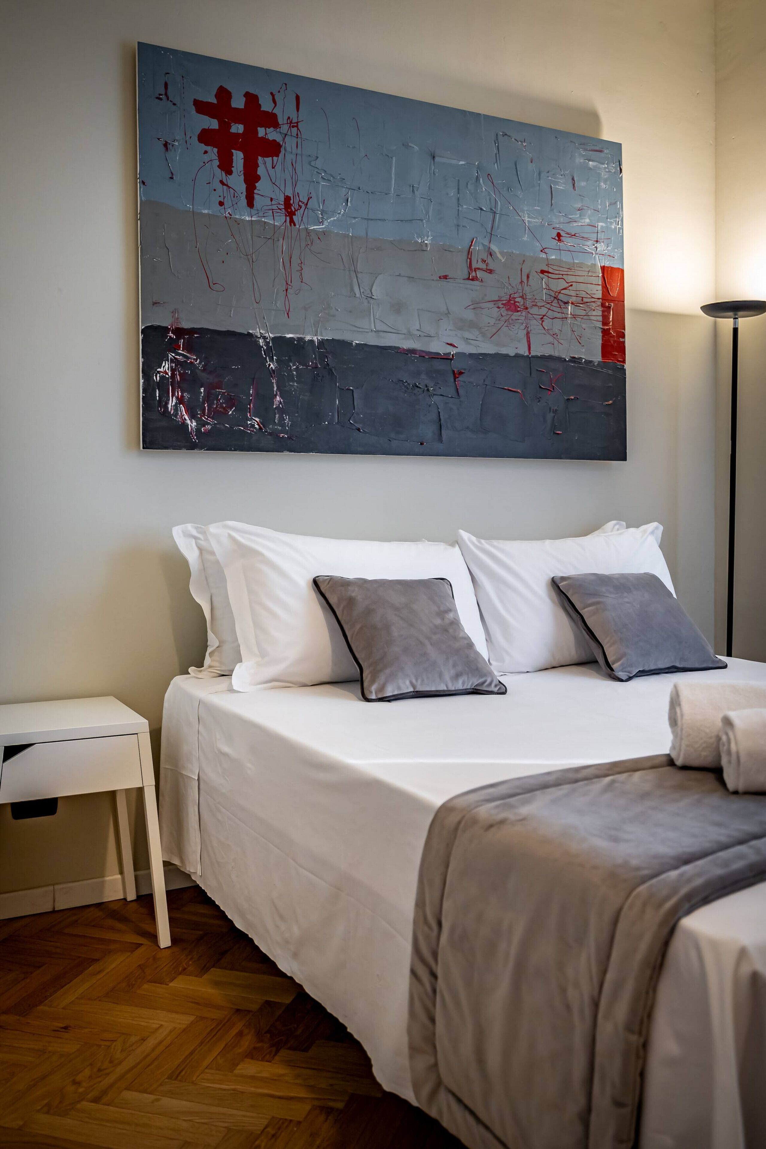 A bedroom with a bed, white pillows, a grey throw blanket, and a modern abstract painting on the wall.