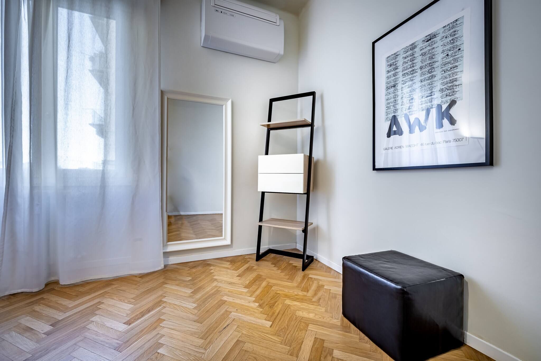 Une pièce dotée d’un parquet en bois à motifs chevrons, d’un pouf en cuir noir, d’une étagère en métal noir et d’une œuvre d’art encadrée accrochée au mur.