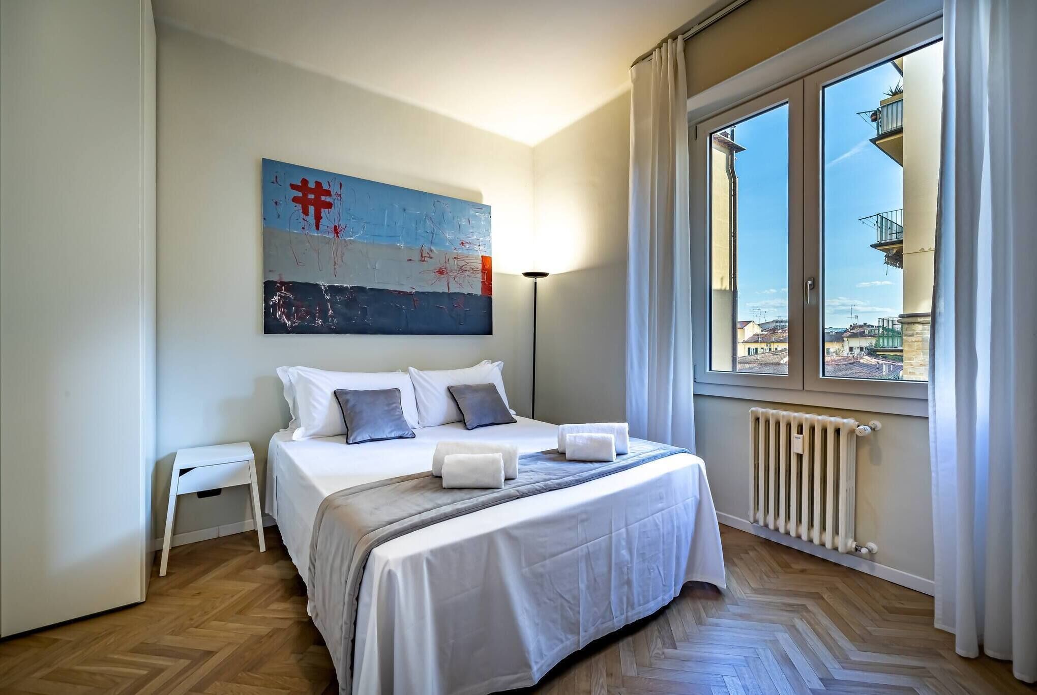 Habitación de hotel con una cama grande, una mesita de noche pequeña, un radiador y una ventana con vistas a edificios.