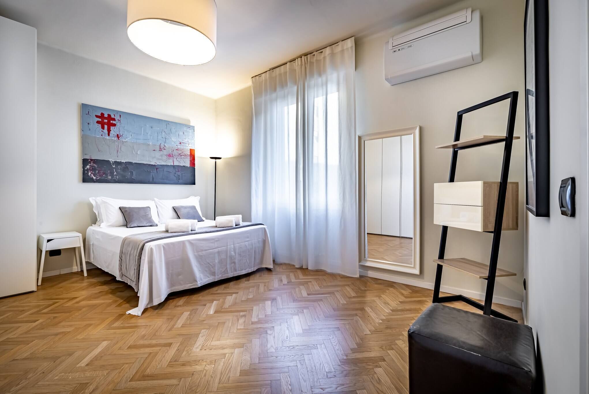 Um quarto moderno com soalho de madeira em padrão herringbone, cama com roupa de cama branca, mesa de cabeceira, candeeiro e um grande quadro abstrato na parede.
