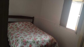 Een kleine kamer met een enkel bed, een bloemenprint bedsprei, een houten hoofdbord en een raam met uitzicht.