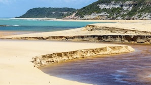 Ein Strand mit klarem Wasser, sandigem Ufer und einer felsigen Klippe im Hintergrund.
