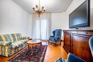 Een woonkamer met een bank, fauteuil, televisie, houten kast en een kroonluchter.