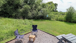 An outdoor seating area with two chairs and a table, surrounded by gravel and grass, with trees in the background. - Sid's Nest (Whitchurch)