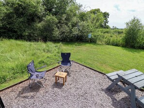 An outdoor seating area with two chairs and a table, surrounded by gravel and grass, with trees in the background. - Sid's Nest (Whitchurch)