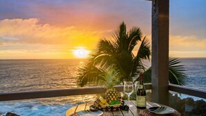 Ein Holztisch mit einer Weinflasche, einer Ananas und einer Obstschale auf einem Balkon mit Blick auf den Ozean bei Sonnenuntergang.