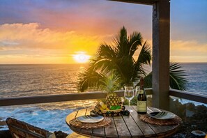 Ein Holztisch mit einer Weinflasche, einer Ananas und einer Obstschale auf einem Balkon mit Blick auf den Ozean bei Sonnenuntergang.