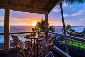 Ein Holztisch mit Stühlen und einer Weinflasche, mit Blick auf einen Sonnenuntergang über dem Meer mit Palmen.
