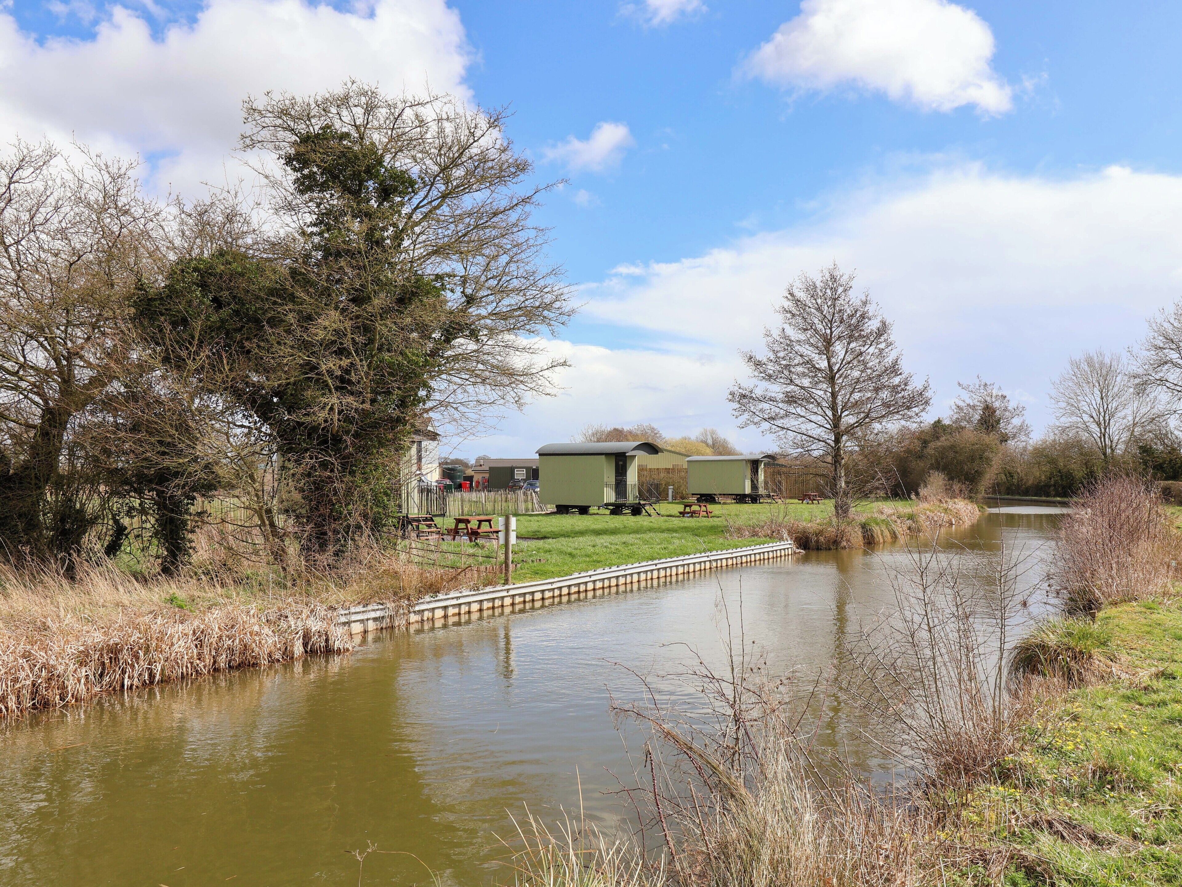 A riverside campsite with green cabins, picnic tables, and a calm water body.