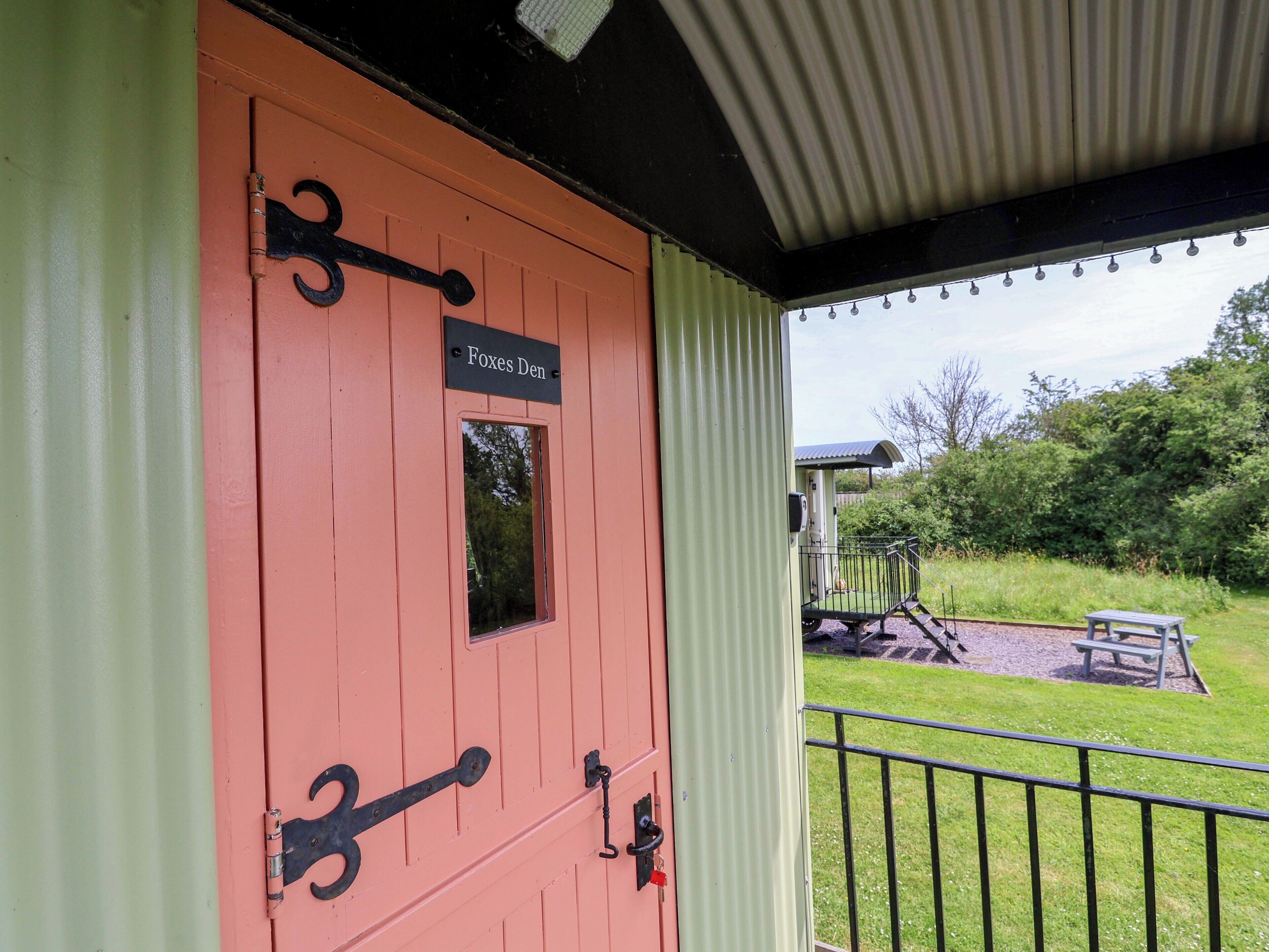 Une porte rose sur laquelle on peut lire « Repaire des renards », une grille en métal et une zone herbeuse avec un banc et une petite construction en arrière-plan.