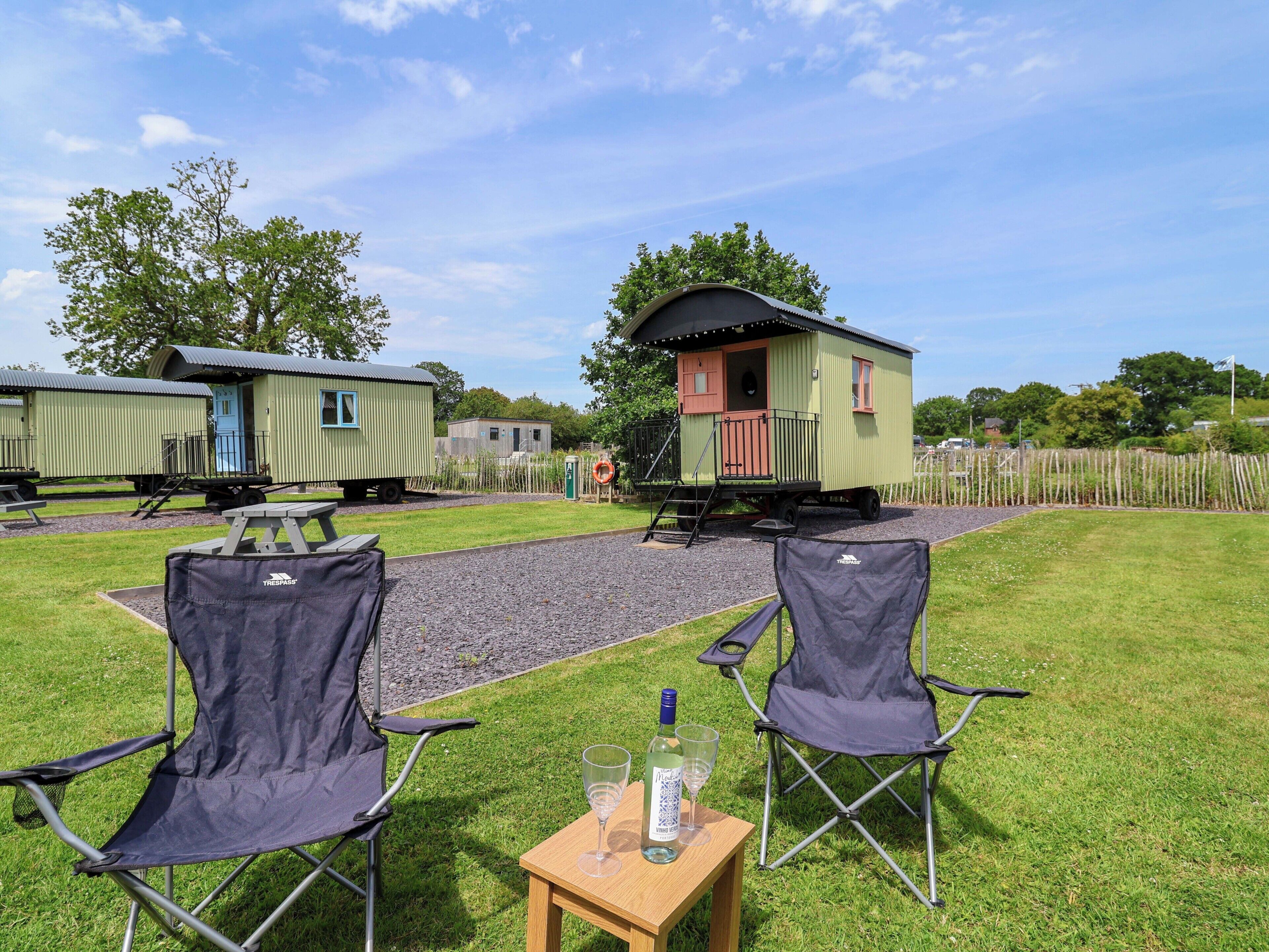 A campsite with two green mobile homes, a gravel path, and two camping chairs.