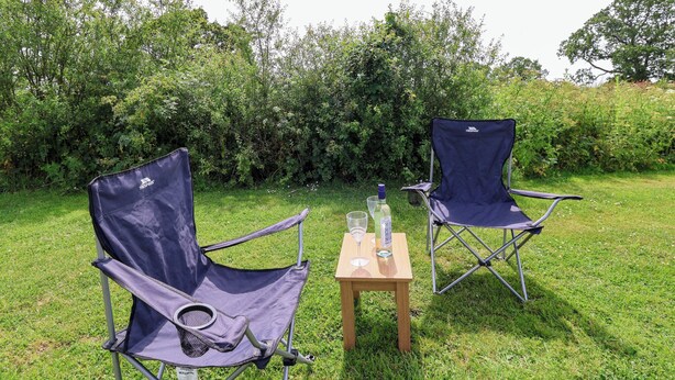 Two camping chairs and a small table with a bottle and glass in a grassy outdoor setting. - Foxes Den (Whitchurch)