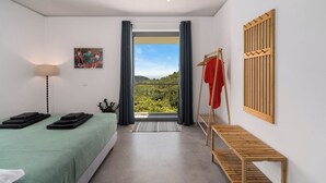 Quarto com cama, mesa de cabeceira, luminária pendente, pintura, janela com cortinas e vista para o verde.